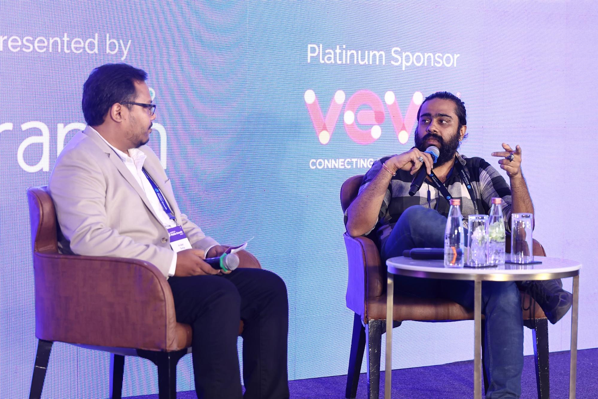 Srinivas Seshadri is seated on stage, speaking into a microphone, addressing another man also seated on stage.